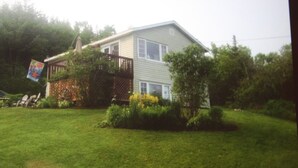 A single-story house with a front deck, surrounded by a well-maintained lawn and trees. - Charming cottage minutes from Gaelic College and Baddeck with WIFI (Baddeck)