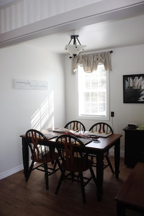 A dining area with a wooden table and chairs, a window with curtains, a wall-mounted light fixture, and a framed quote on the wall.