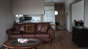 A living room with a brown leather sofa, a glass coffee table, a kitchen area with a refrigerator and microwave, and a dining area with a table and chairs. - Charming cottage minutes from Gaelic College and Baddeck with WIFI (Baddeck)