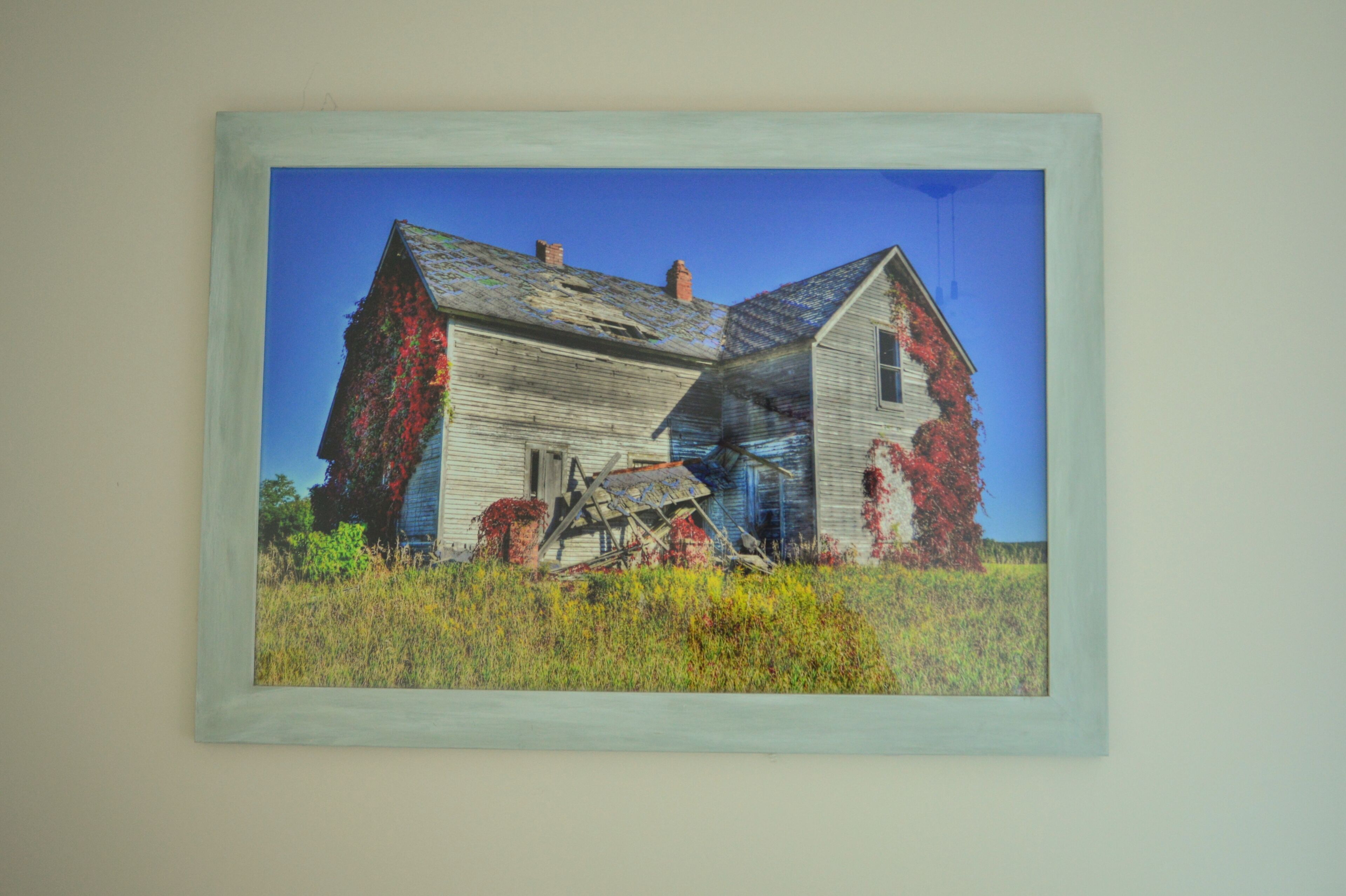 A painting of a dilapidated house with overgrown vegetation and a clear blue sky.