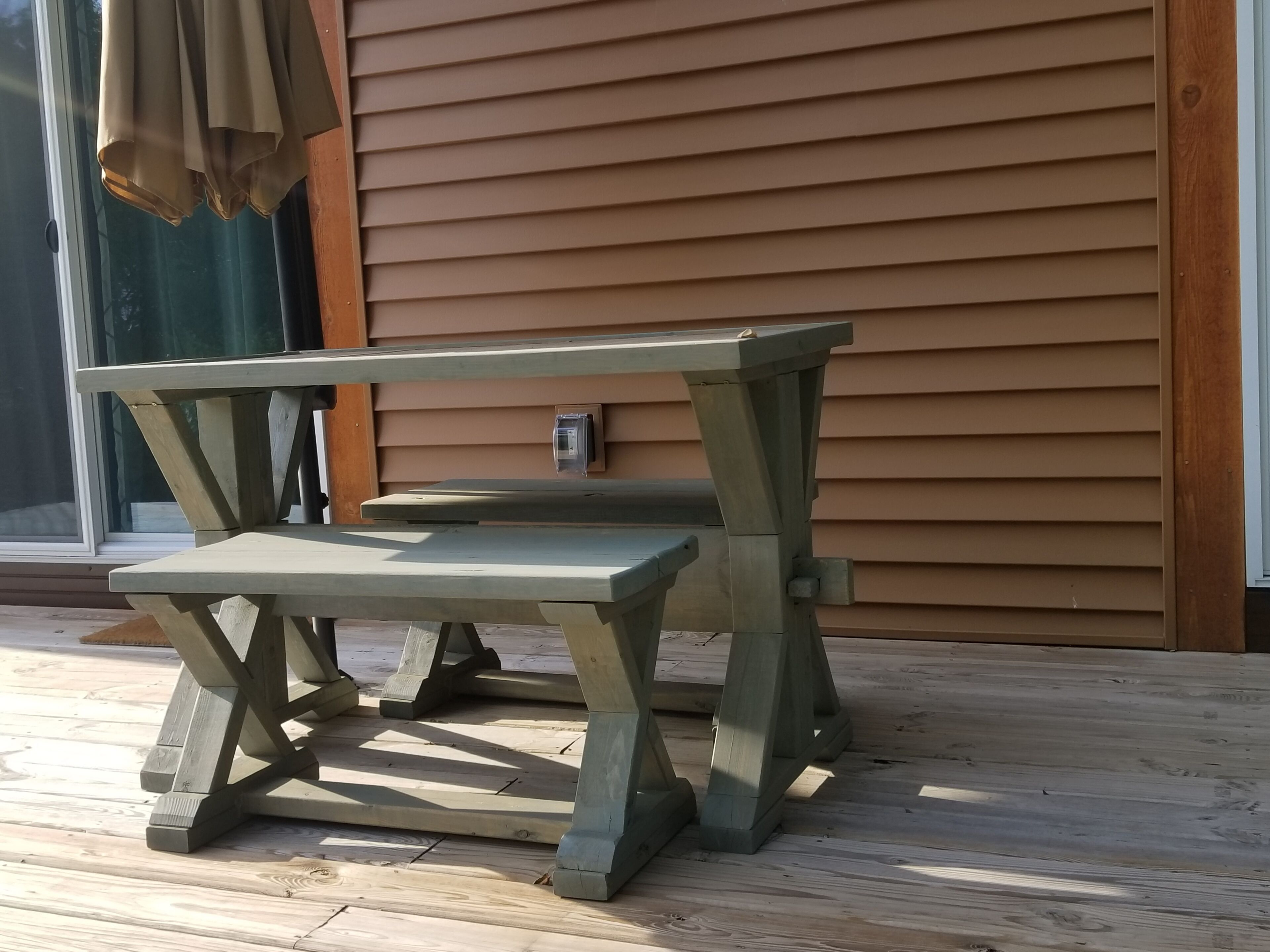A wooden picnic table with a bench, positioned on a wooden deck in front of a house with horizontal siding.