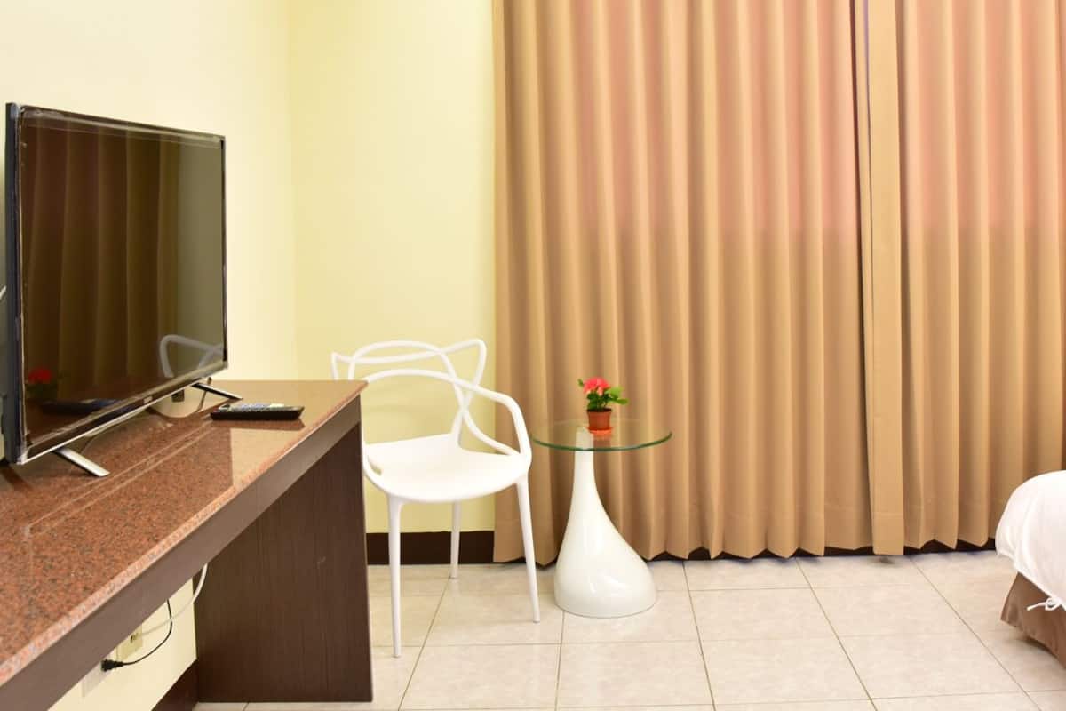Hotellihuone, jossa on sänky, työpöytä, jossa on televisio, tuoli, pieni pöytä maljakolla ja verhot.