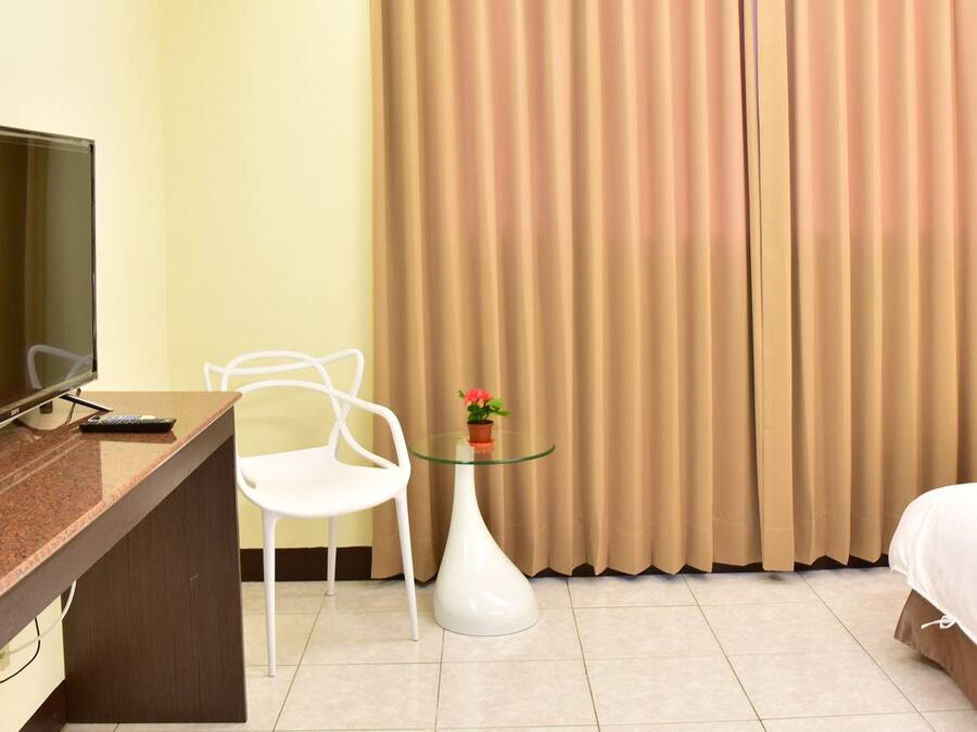 A hotel room with a bed, a desk with a television, a chair, a small table with a vase, and curtains.