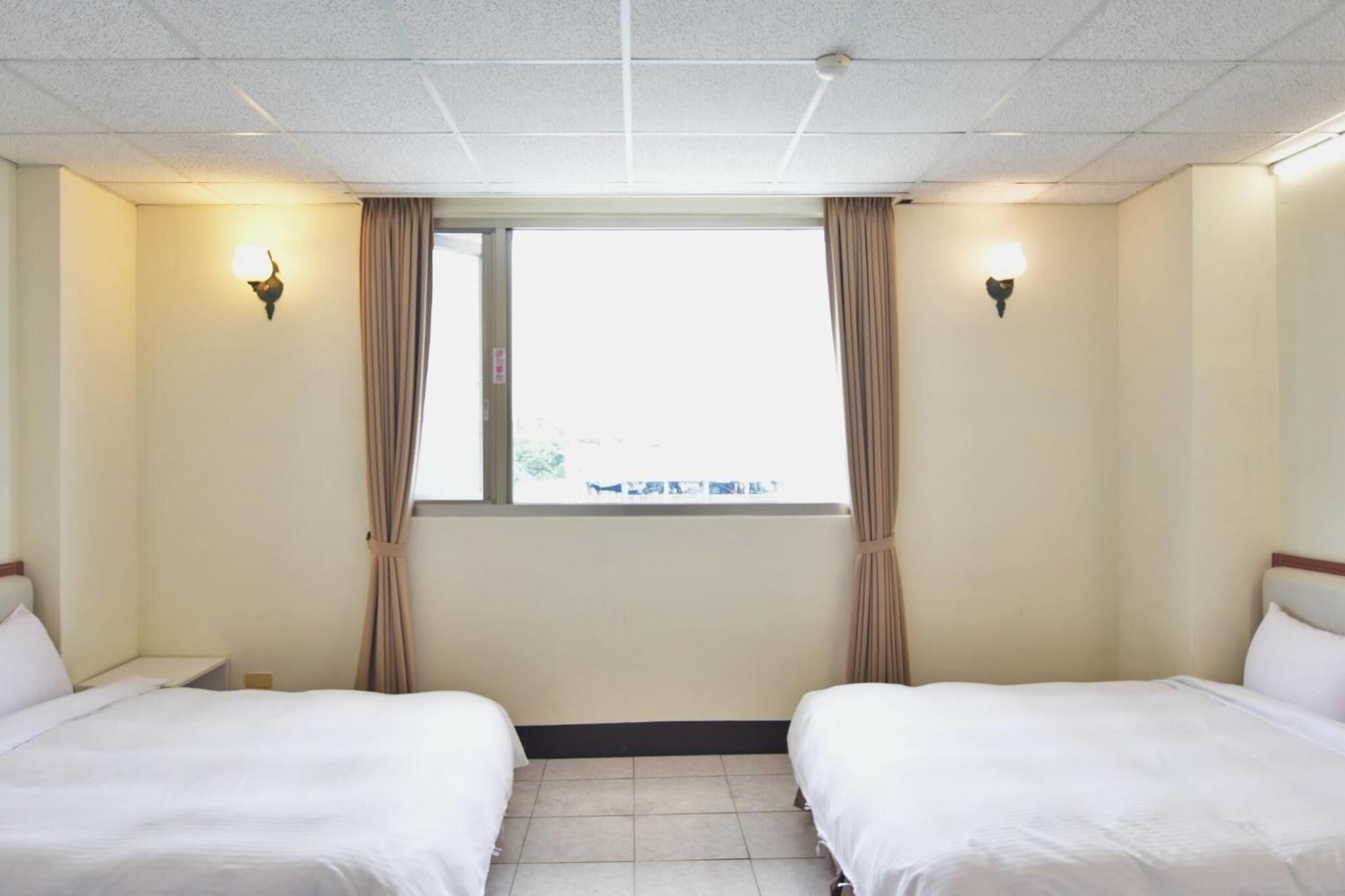 A hotel room with two beds, a window with curtains, and wall-mounted lights.