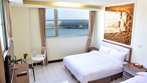 A hotel room with a bed, a desk with a remote control, a chair, and a window with a view of the sea. - Fugong Hostel (Taitung)