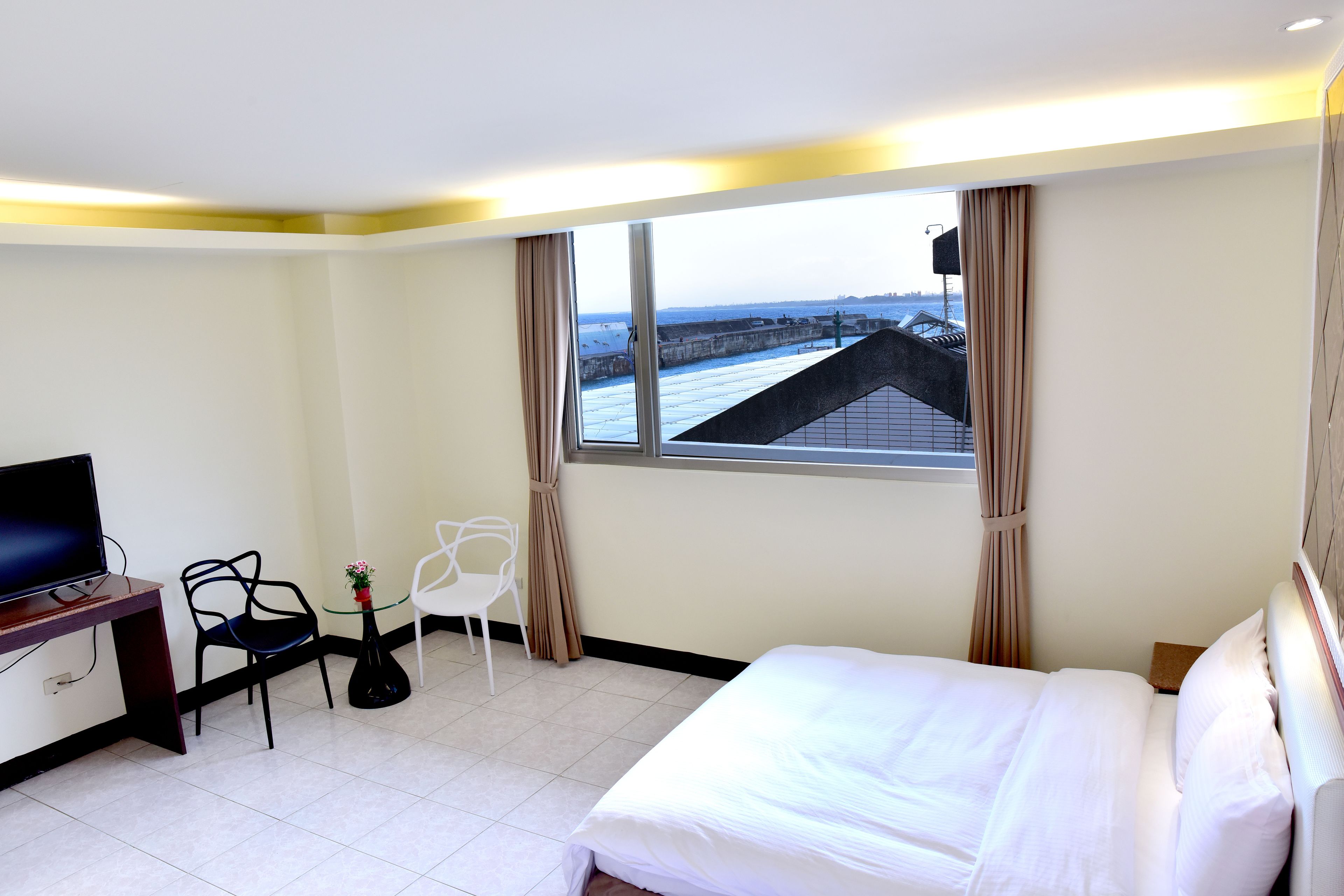 Habitación de hotel con cama, escritorio con televisor, silla, pequeña mesa con planta y ventana con vista a un puente.
