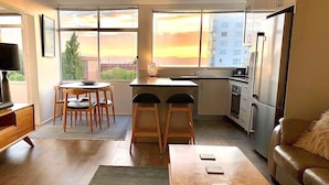Cozinha moderna com área de refeições, sofá e vista do pôr do sol por grandes janelas.