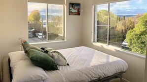 Quarto com cama, almofadas e janela com vista para árvores e edifícios.