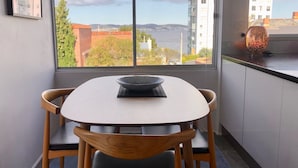 Uma área de refeições com mesa e cadeiras, vista para um corpo de água e edifícios, e um objeto decorativo no parapeito da janela.