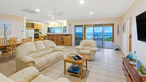 A spacious living room with a sofa, armchairs, and a coffee table. There is a kitchen area with cabinets and a dining table. The room has large windows with a view of the ocean. - 'IslandView' The Perfect Holiday Home (Hideaway Bay)