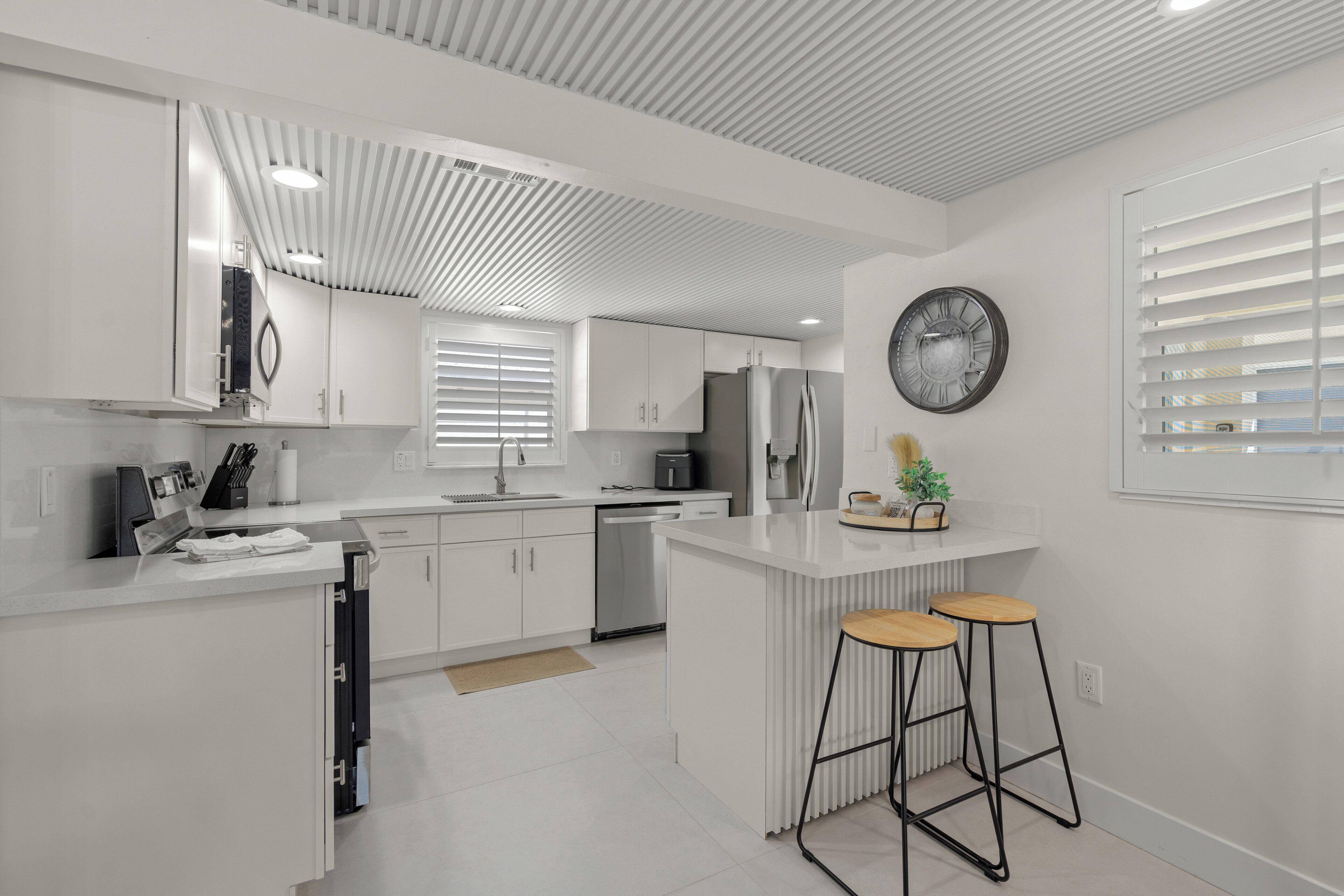 Una cocina moderna con armarios blancos, una isla central con taburetes, un horno empotrado y un reloj en la pared.