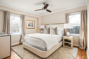 Quarto com cama, duas mesas de cabeceira, ventilador de teto, janela com cortinas e um quadro na parede.