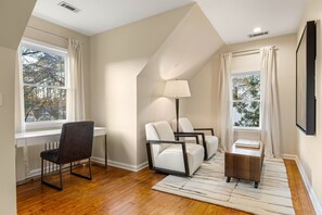 A modern living room with a sloped ceiling, a desk with a chair, two sofas, a floor lamp, and a TV mounted on the wall.