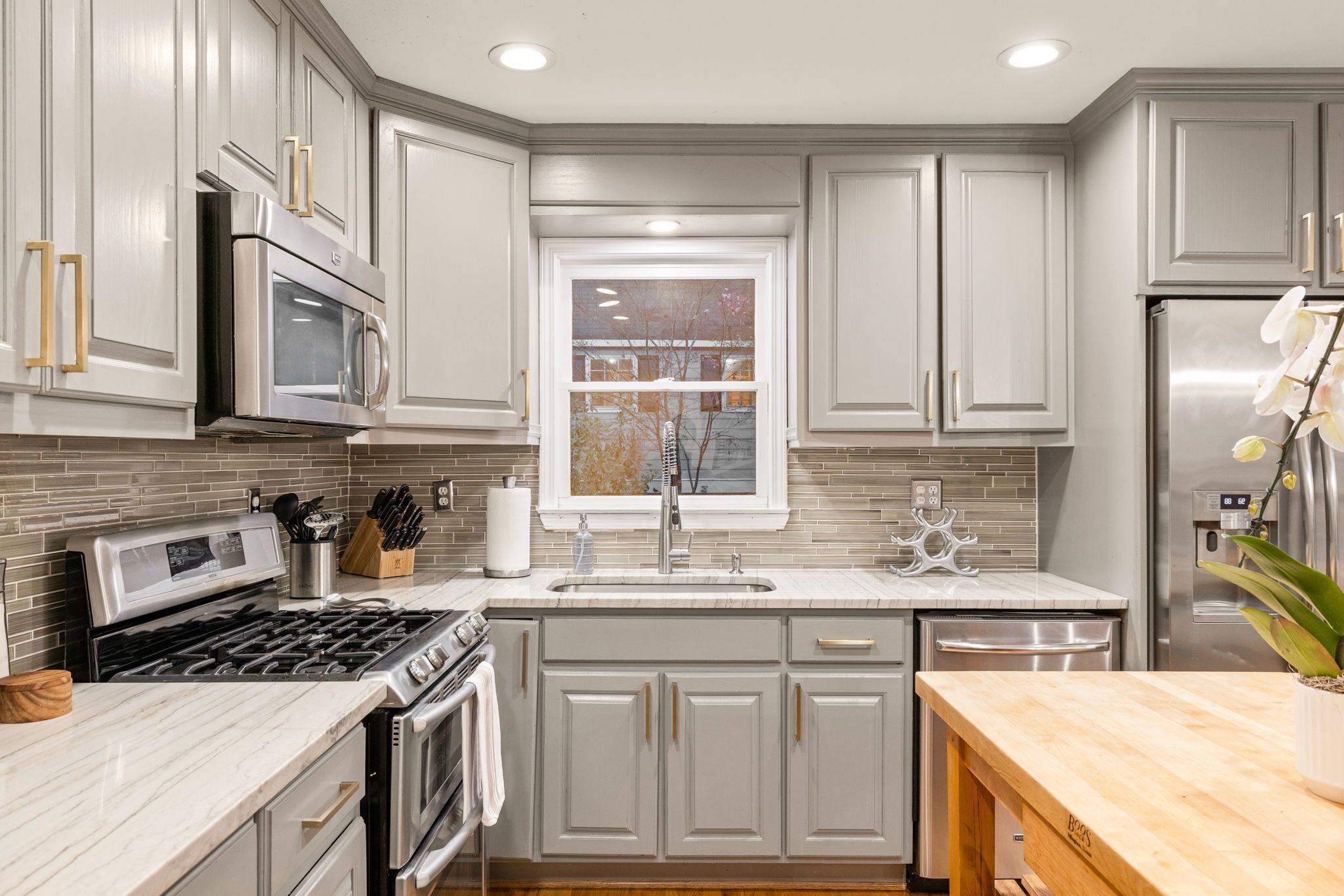 A modern kitchen with grey cabinetry, stainless steel appliances, a marble countertop, and a window with a view of trees.