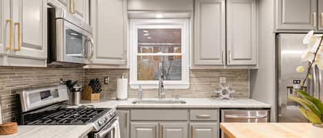 A modern kitchen with grey cabinetry, stainless steel appliances, a marble countertop, and a window with a view of trees.