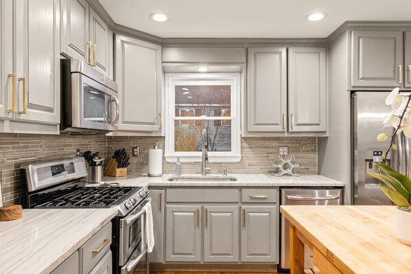 A modern kitchen with grey cabinetry, stainless steel appliances, a marble countertop, and a window with a view of trees.