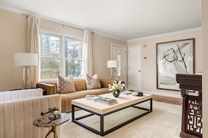 A living room with a sofa, coffee table, and a lamp. There is a door and a window with curtains.