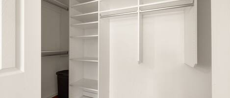 A walk-in closet with multiple shelves and hanging space, wooden flooring, and a trash can.