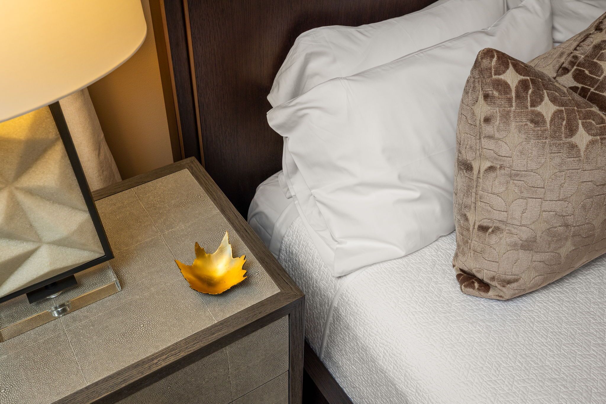 Un lit bien fait, avec du linge de lit blanc et un seul oreiller marron, une petite table de chevet avec une lampe et un objet décoratif doré.