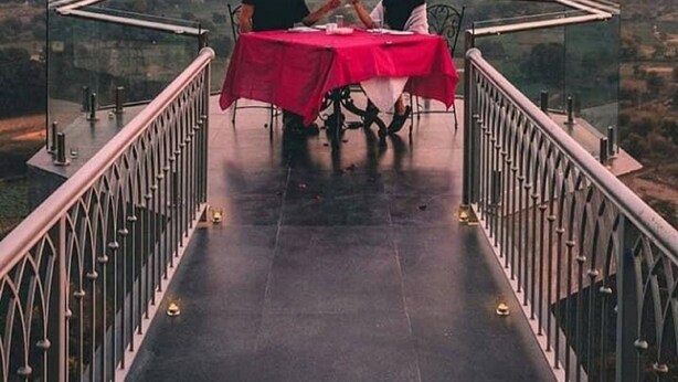 A table with a red tablecloth on a balcony with glass railings, overlooking a cityscape and mountains. - Fateh Niwas (Udaipur)