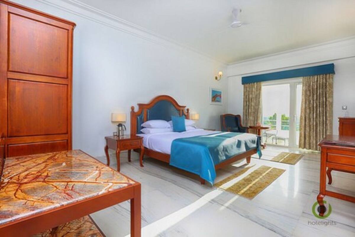 A hotel room with a bed, wooden furniture, a table, and a view of greenery outside.