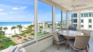 Un balcon avec une table en verre et des chaises en osier, offrant une vue sur la plage et le paysage urbain.