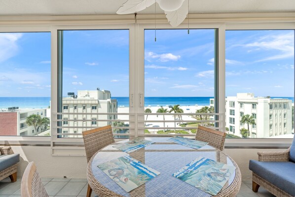Un balcón con mobiliario de mimbre, una mesa redonda con vistas a la playa y ventanas amplias con vista al océano.