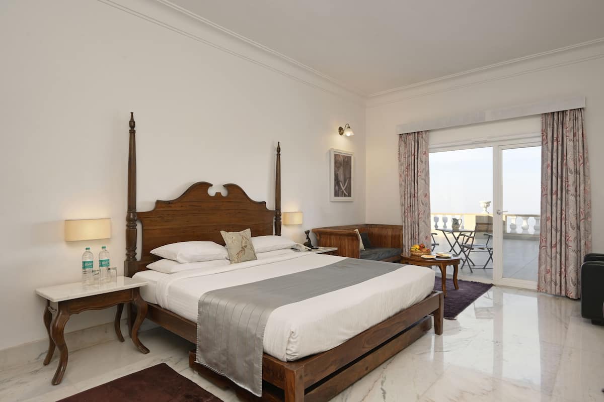 A hotel room with a large bed, bedside tables, a small table with a chair, a balcony with curtains, and a view of the outside.