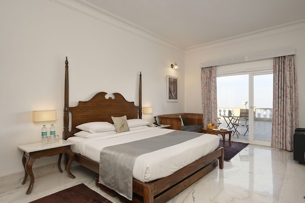 Habitación de hotel con una cama grande, mesitas de noche, una pequeña mesa con una silla, un balcón con cortinas y vistas al exterior.