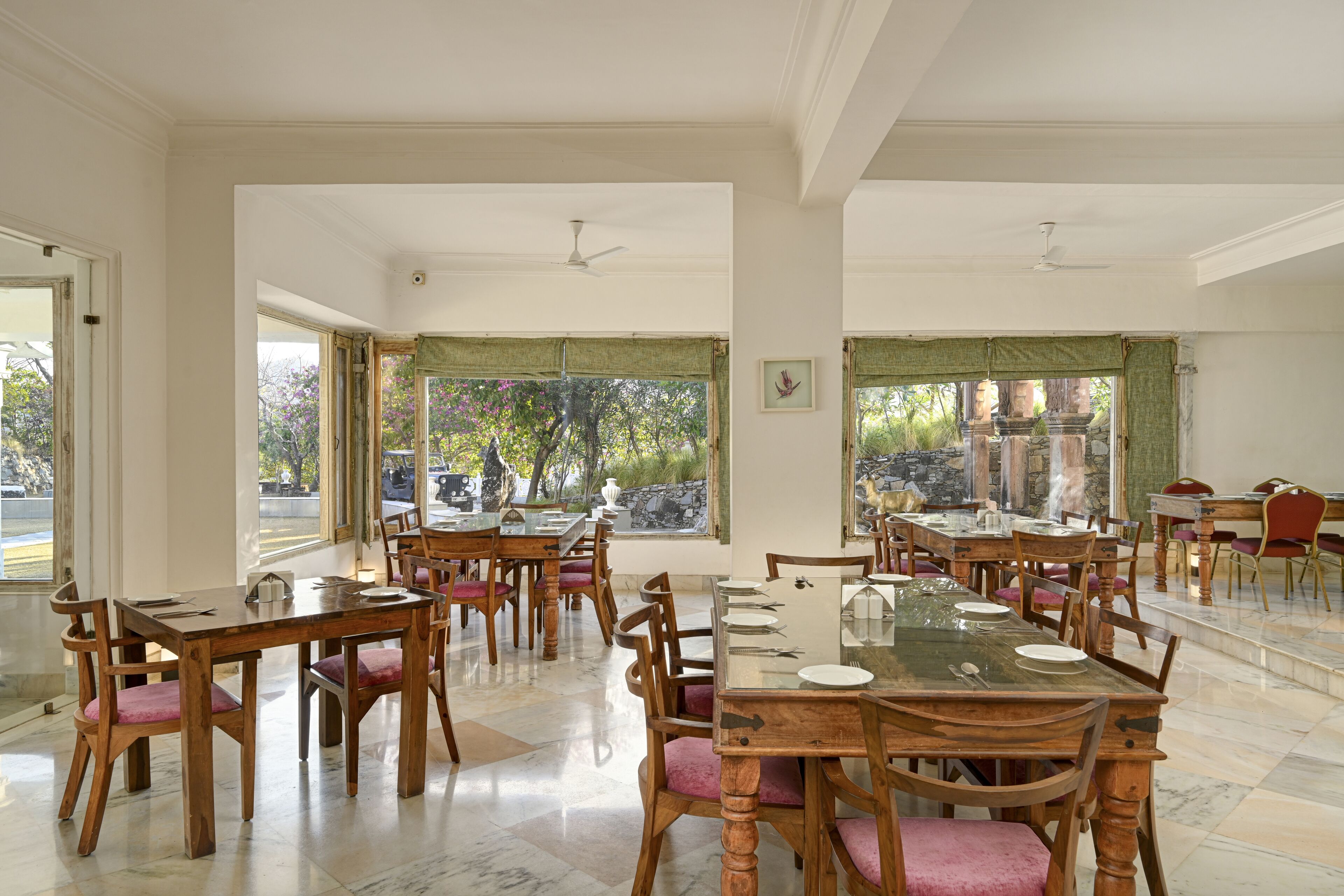 A spacious dining area with multiple wooden tables and chairs, large windows with green curtains, and a view of trees and buildings outside.