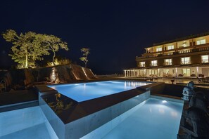 A swimming pool with a stone border, a stone statue, and a building with multiple balconies and lights. - Fateh Safari Resort by Fateh Collection (Kumbhalgarh)