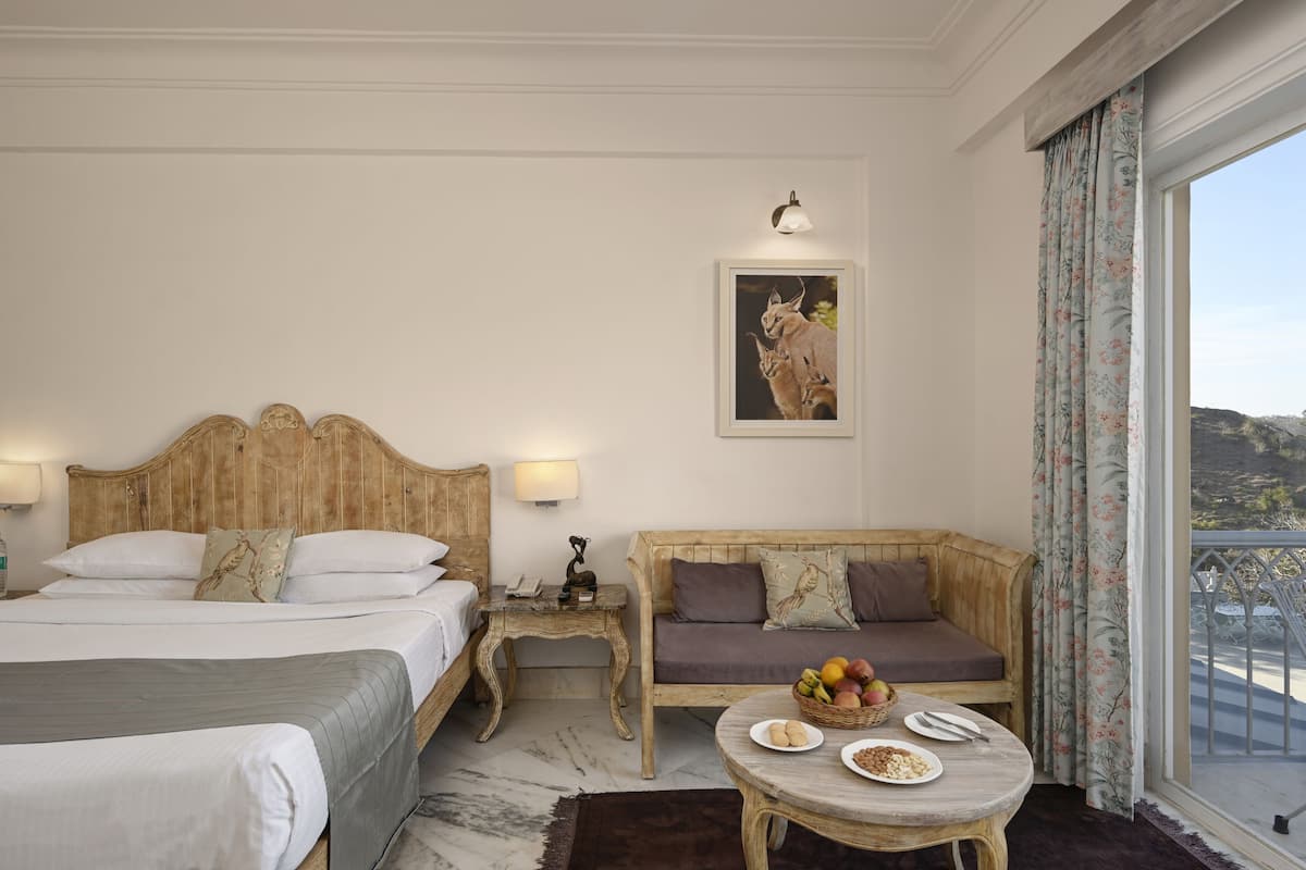 A hotel room with two beds, a sofa, a dining table with plates and a fruit basket, a lamp, and a framed picture on the wall.