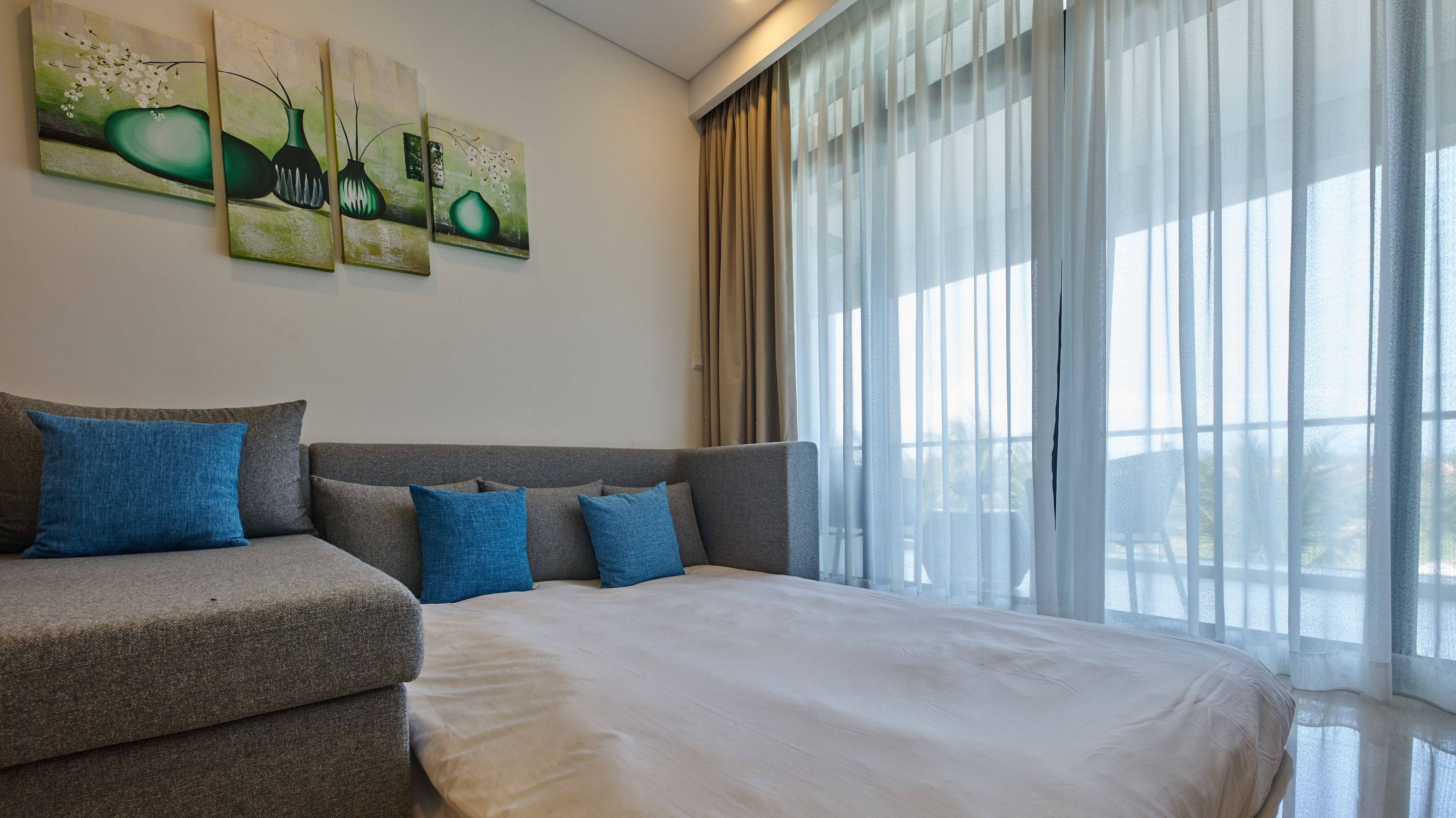 Une chambre d’hôtel moderne avec un grand lit, un canapé gris agrémenté de coussins bleus et trois tableaux accrochés au mur.