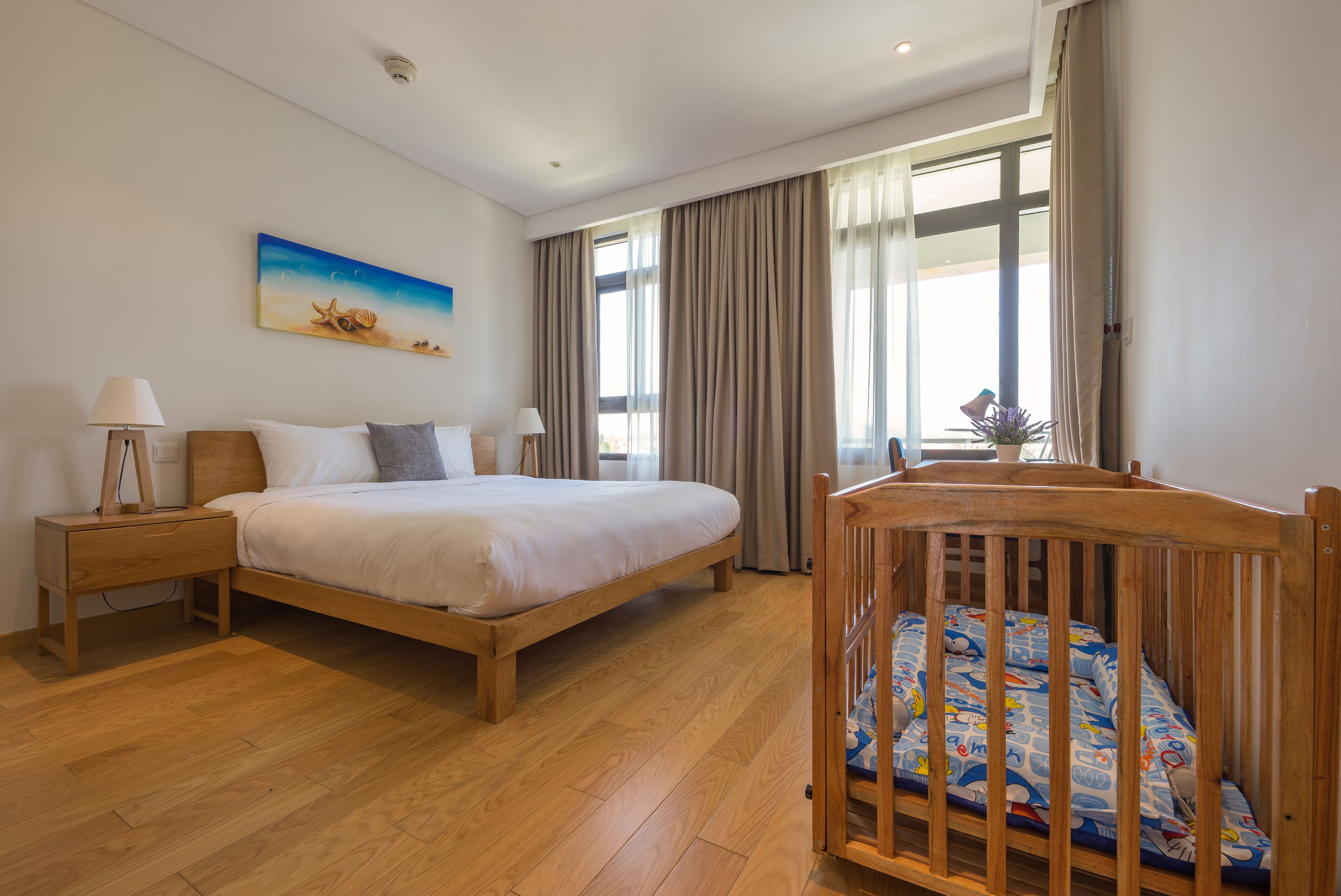 Un dormitorio con cama de madera, mesita de noche, cuna, un cuadro de una playa y ventanas amplias con cortinas.