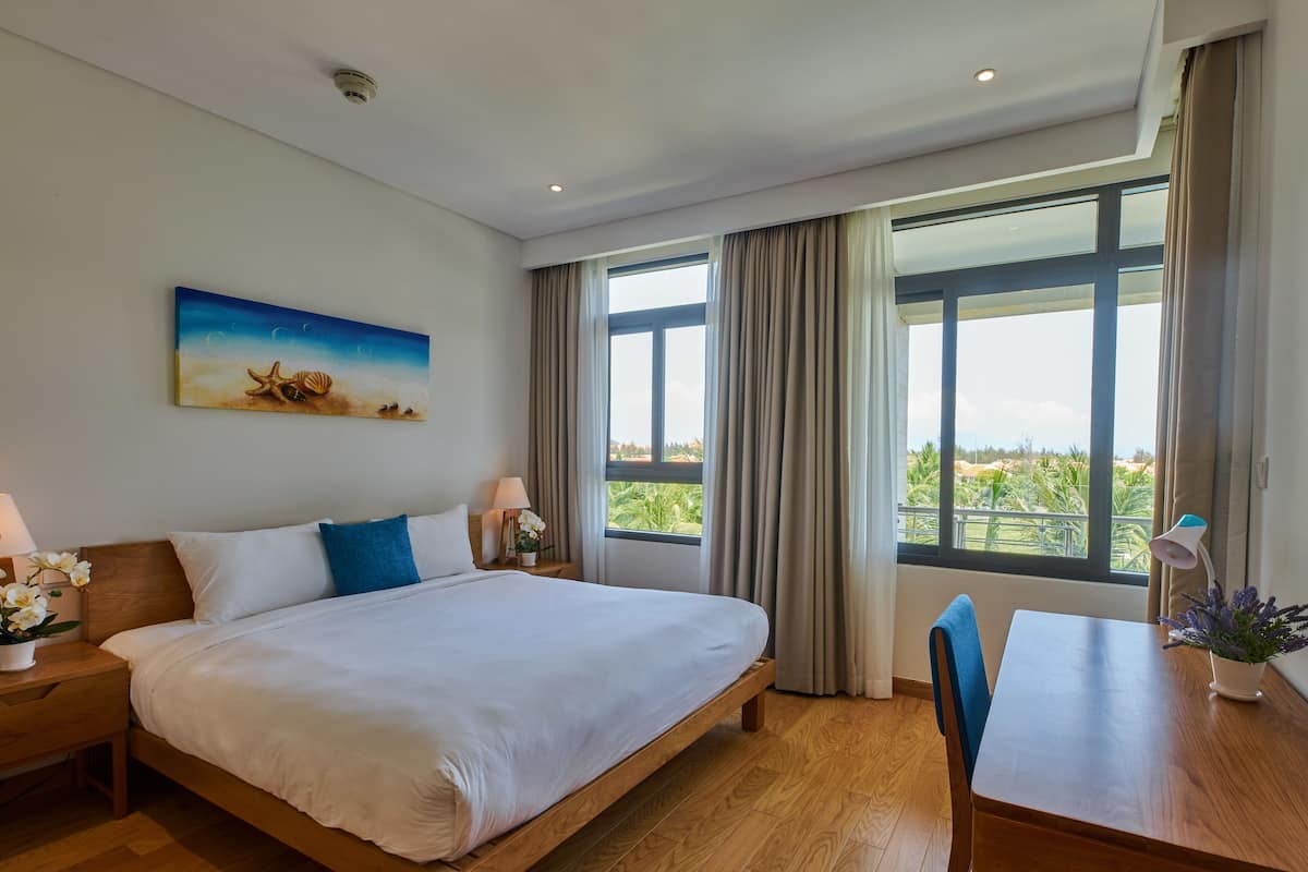 A hotel room with a large bed, a desk with a chair, a painting on the wall, and a view of the outdoors through the windows.