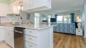 A modern kitchen with white cabinets, a dishwasher, and a large island. There is a dining area with a striped sofa and a lamp.