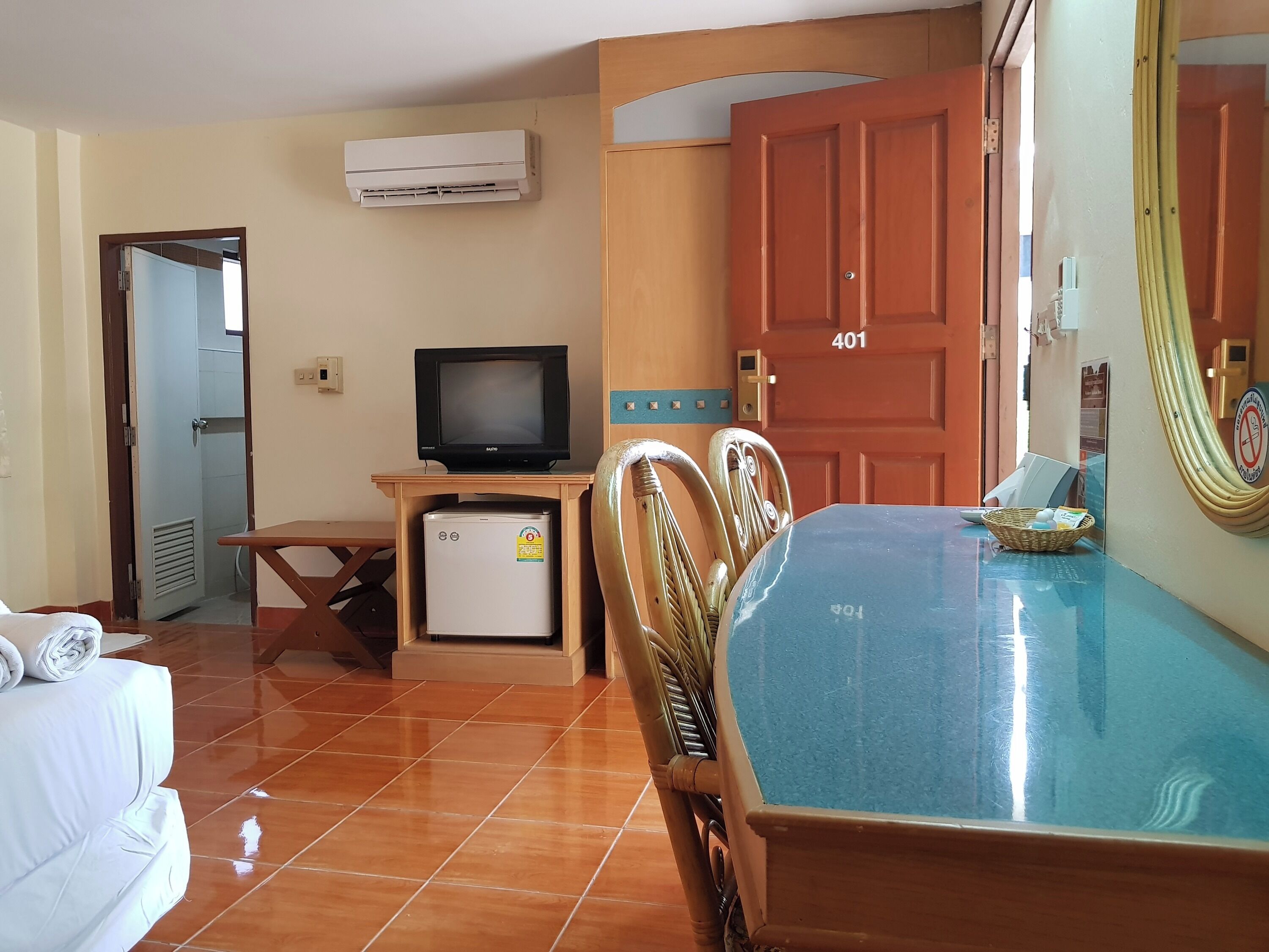 Una habitación con cama, una mesa, sillas, televisión, refrigerador pequeño y aire acondicionado, además de una puerta con un número.