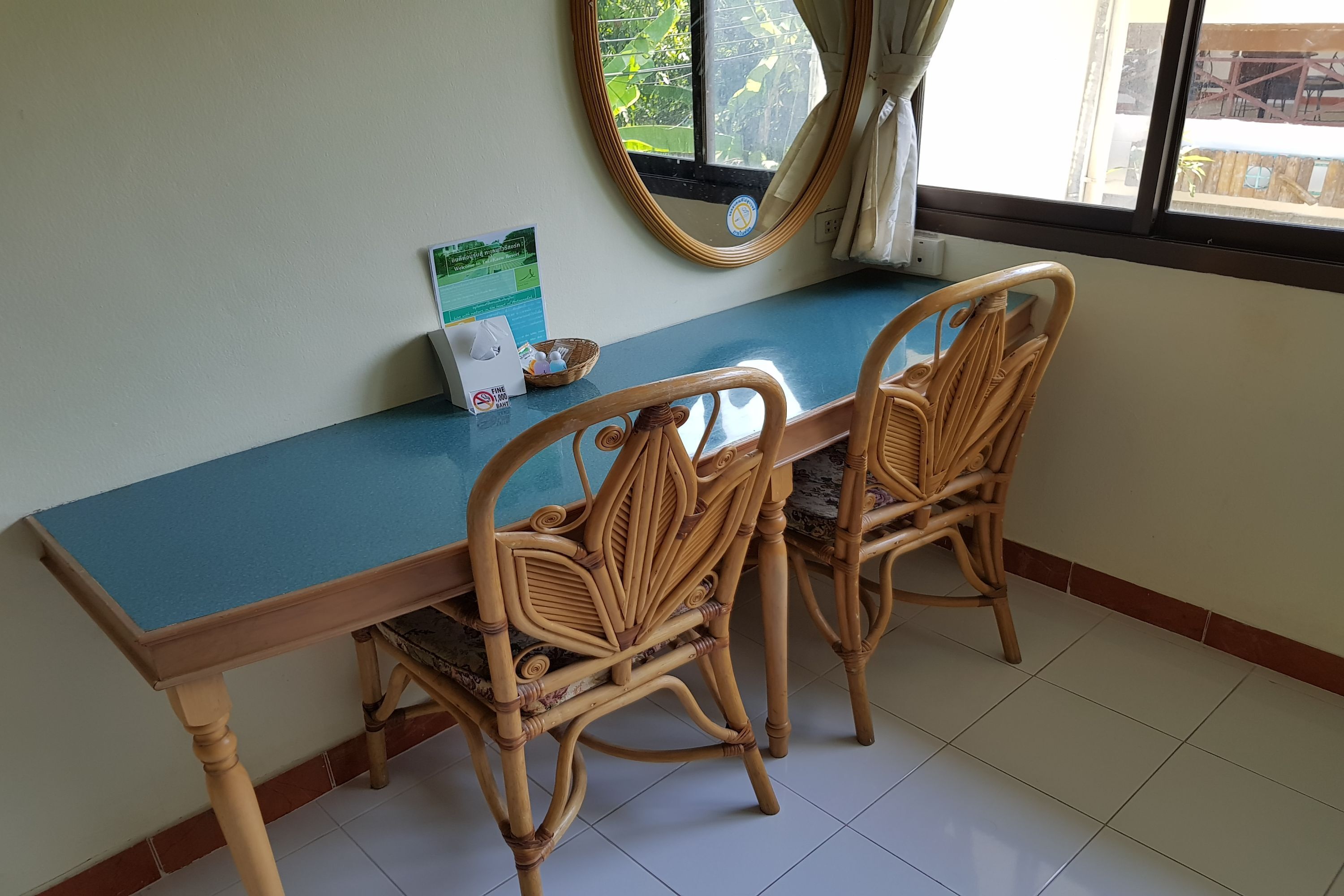 A dining area with a blue table and two wicker chairs, a mirror on the wall, and a window with a view of greenery outside.