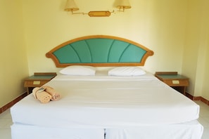 Una cama bien hecha con sábanas blancas y un cabecero tapizado en color verde mar. Hay dos mesitas de noche con lámparas, y un par de toallas enrolladas sobre la cama.