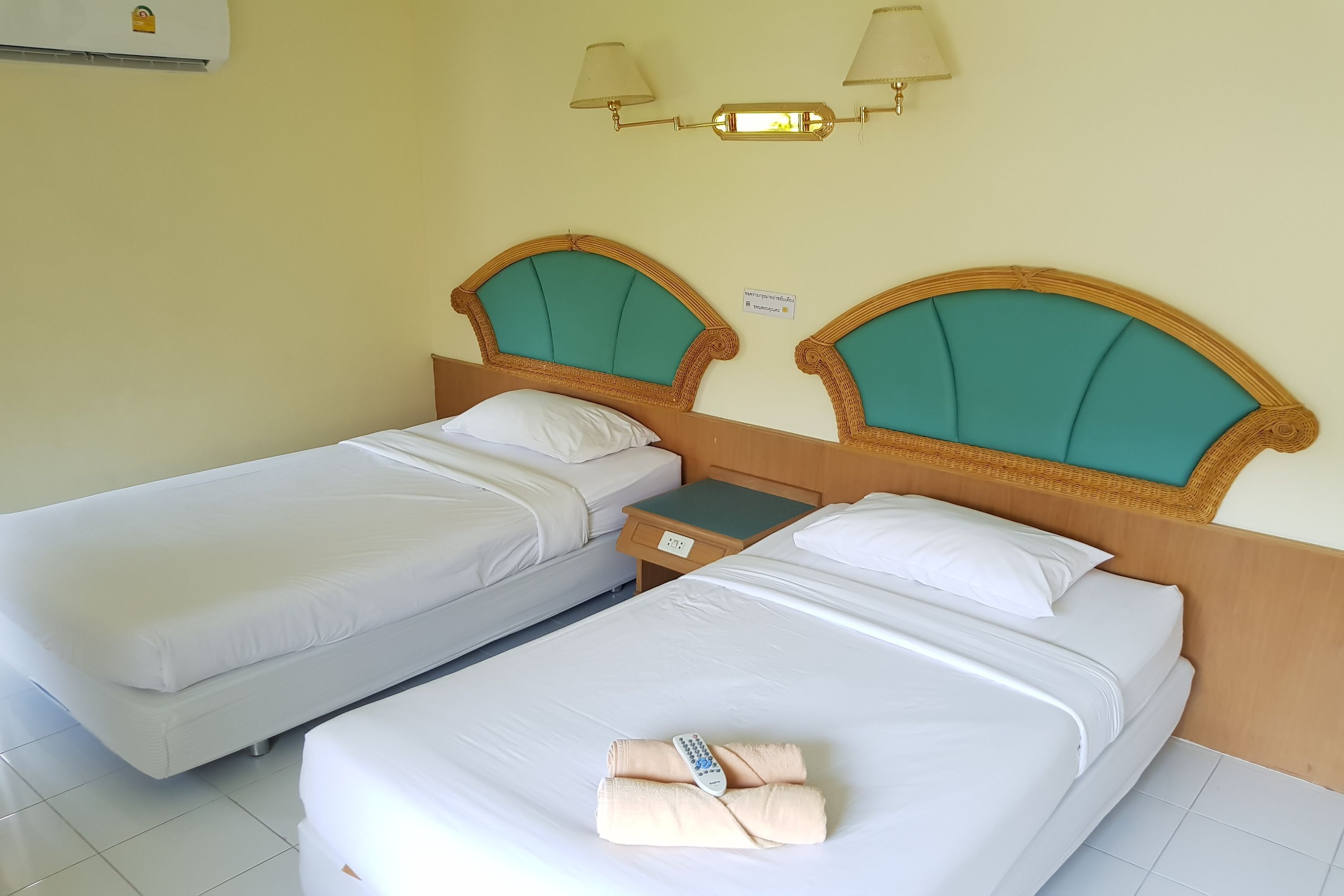 A hotel room with two single beds, each with a wooden headboard and teal cushions. There is a nightstand with a lamp and a small table between the beds.