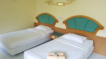 A hotel room with two single beds, each with a wooden headboard and teal cushions. There is a nightstand with a lamp and a small table between the beds.
