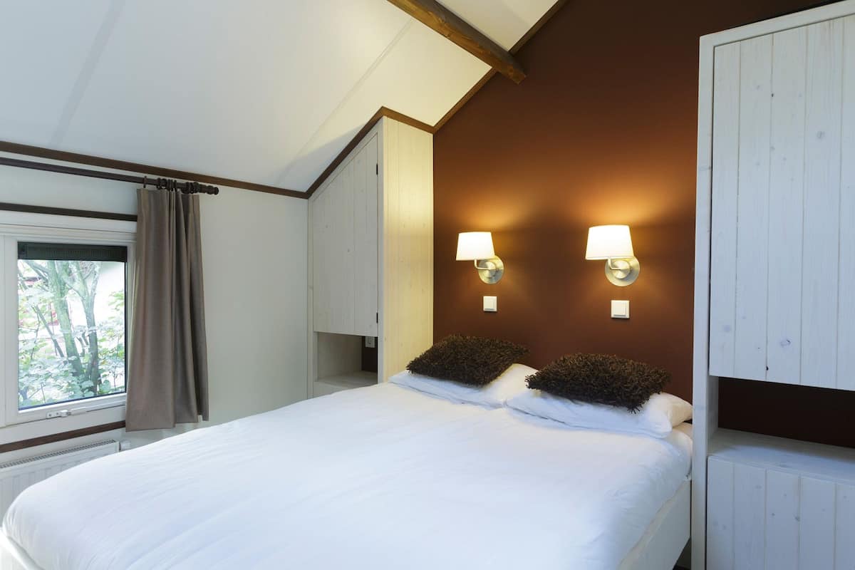 A bedroom with a large bed, two wall-mounted lamps, a window with curtains, and a wooden ceiling.