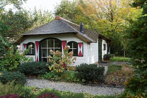 A small, white cottage with a thatched roof, surrounded by lush greenery and flowering plants. - Vrijrijck Vakantieparken – Ermelo (Ermelo)
