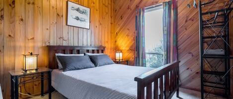 Quarto de cabana de madeira com uma cama, mesas de cabeceira, uma luminária pendente e uma janela com cortinas.