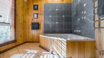 Ein Badezimmer mit Holzwandverkleidung, einer freistehenden Badewanne, gefliesten Wänden und einem Fenster mit Jalousien.