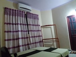 A room with a bed, an air conditioner, a window with purple curtains, and a door.