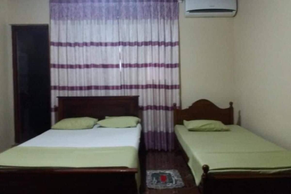 A room with two single beds, a wooden headboard, a curtain with horizontal stripes, and an air conditioning unit on the wall.