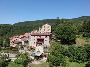 A traditional stone building with multiple levels and a red-tiled roof, surrounded by lush greenery and hills. - Alla Battuta (Sansepolcro)