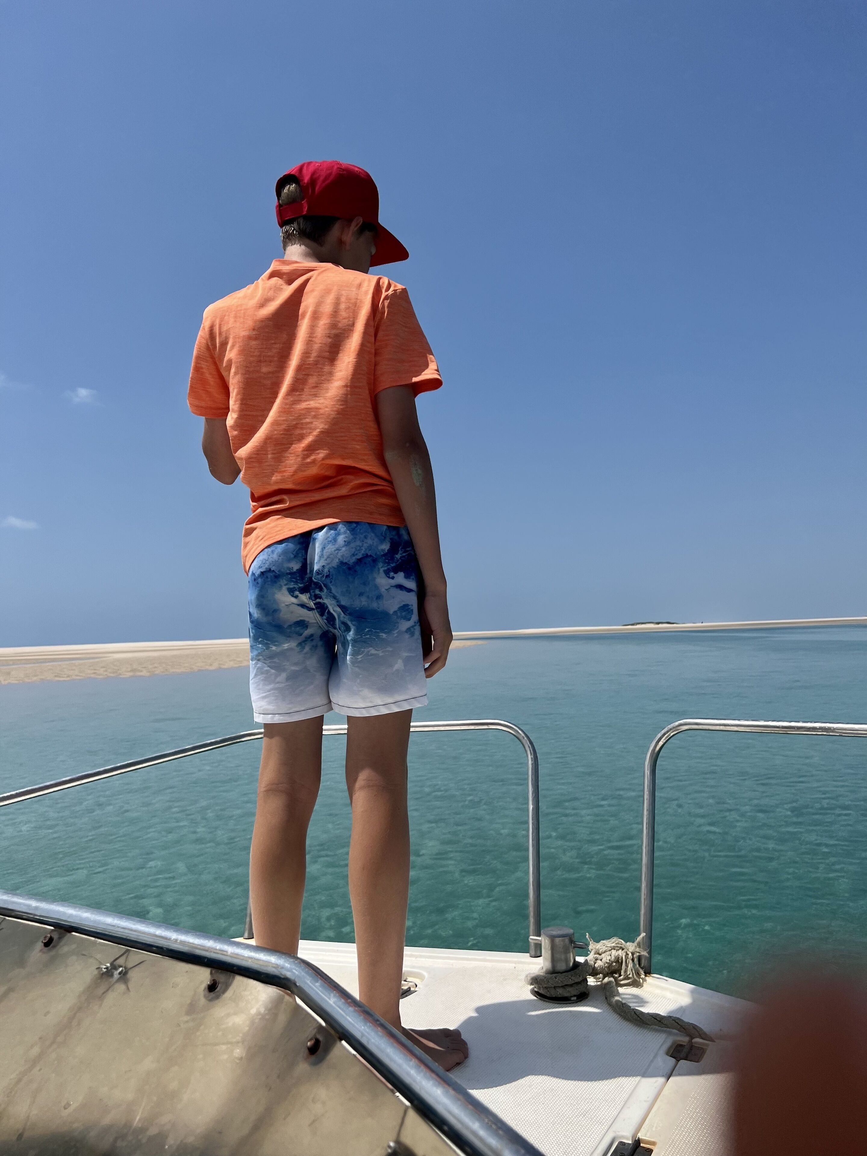 Une personne se tient sur le pont d’un bateau, surplombant une mer calme, avec une plage de sable qui s’étend au loin.