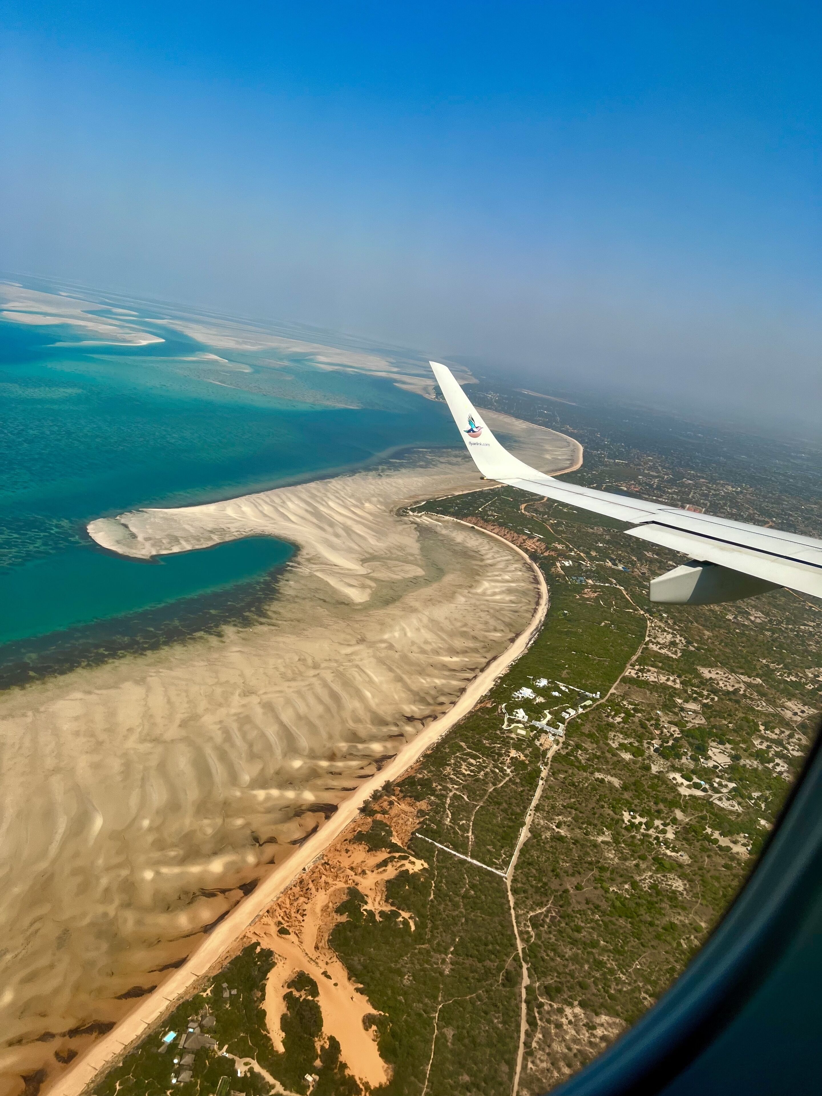 Vista aérea de una zona costera con playa de arena, aguas azules cristalinas y una combinación de vegetación verde y desarrollo urbano.